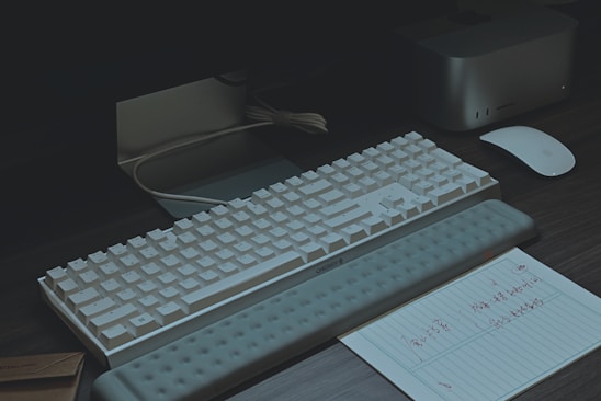 A modern workspace setup featuring a white mechanical keyboard with a wrist rest, an Apple Magic Mouse, and a note with handwritten text on lined paper. There is a mini PC or docking station nearby, along with a monitor. The lighting is dim, creating a moody atmosphere.