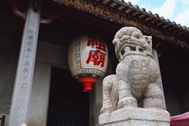 A stone lion statue stands prominently in front of a traditional building entrance. Above the statue, a large lantern with red Chinese characters hangs from the ceiling. The entrance is framed with carved stone columns featuring vertical inscriptions.