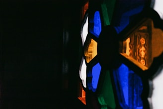 A close-up of a stained glass window featuring vibrant geometric patterns and colors. The design includes various shapes with rich hues of blue, white, green, yellow, and orange against a dark background.