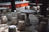 Stone tables and stools are arranged in an outdoor setting, surrounded by trees. The setting is shaded, with sunlight filtering through the leaves creating patterns on the ground. The furniture has a rustic and traditional design, likely made of granite or a similar material.