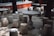 Stone tables and stools are arranged in an outdoor setting, surrounded by trees. The setting is shaded, with sunlight filtering through the leaves creating patterns on the ground. The furniture has a rustic and traditional design, likely made of granite or a similar material.
