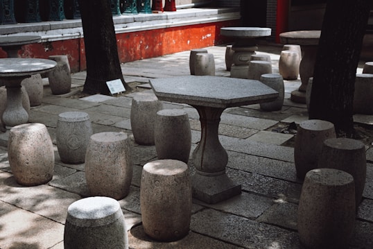Stone tables and stools are arranged in an outdoor setting, surrounded by trees. The setting is shaded, with sunlight filtering through the leaves creating patterns on the ground. The furniture has a rustic and traditional design, likely made of granite or a similar material.