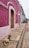 A rustic stone walkway leads along a vibrant pink and white building with wooden doors. A dog rests on the path, enjoying the peaceful surroundings. In the distance, a quaint outdoor seating area with green umbrellas is visible, alongside colonial-style street lamps.
