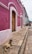 A rustic stone walkway leads along a vibrant pink and white building with wooden doors. A dog rests on the path, enjoying the peaceful surroundings. In the distance, a quaint outdoor seating area with green umbrellas is visible, alongside colonial-style street lamps.