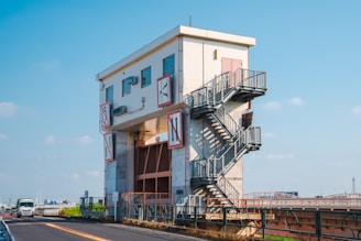 A multi-story industrial building with external metal stairs is positioned beside a road. The structure features signage with symbols and letters. A truck is seen driving on the road, and the background includes a clear blue sky.