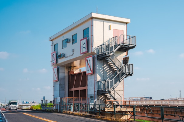 A multi-story industrial building with external metal stairs is positioned beside a road. The structure features signage with symbols and letters. A truck is seen driving on the road, and the background includes a clear blue sky.