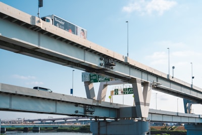 An elevated highway structure with multiple layers of roads and visible vehicles, including a truck with an advertisement and a car. Signage provides directions, and the background shows a clear blue sky with a few clouds.