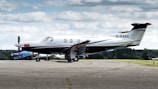 A sleek, single-engine aircraft is parked on an airport tarmac. The plane is predominantly white with black and silver accents, and the tail number G-KARE is visible. In the background, there's another smaller airplane covered with a blue tarp, and a green landscape under a sky filled with scattered clouds.