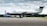 A sleek, single-engine aircraft is parked on an airport tarmac. The plane is predominantly white with black and silver accents, and the tail number G-KARE is visible. In the background, there's another smaller airplane covered with a blue tarp, and a green landscape under a sky filled with scattered clouds.