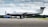 A sleek, single-engine aircraft is parked on an airport tarmac. The plane is predominantly white with black and silver accents, and the tail number G-KARE is visible. In the background, there's another smaller airplane covered with a blue tarp, and a green landscape under a sky filled with scattered clouds.
