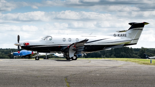 A sleek, single-engine aircraft is parked on an airport tarmac. The plane is predominantly white with black and silver accents, and the tail number G-KARE is visible. In the background, there's another smaller airplane covered with a blue tarp, and a green landscape under a sky filled with scattered clouds.