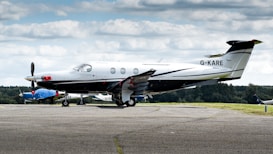 A sleek, single-engine aircraft is parked on an airport tarmac. The plane is predominantly white with black and silver accents, and the tail number G-KARE is visible. In the background, there's another smaller airplane covered with a blue tarp, and a green landscape under a sky filled with scattered clouds.