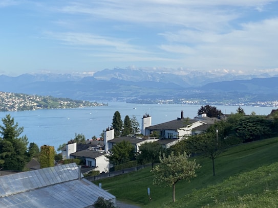 A serene landscape featuring a large lake with gentle waves, surrounded by lush greenery and distant mountains under a partly cloudy sky. Houses with modern architecture are nestled along the hillside, overlooking the water. The overall setting is peaceful and picturesque.
