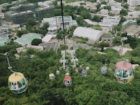 A bird's-eye view of a cable car system with colorful gondolas traveling over a lush green forest. The background features an amusement park with various buildings, attractions, and structures, including a domed building and tennis courts. The image also shows urban buildings in the distance.