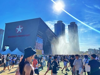 A large outdoor event with many people gathering in front of a stage. The stage has a big screen and is part of a structure labeled 'INCHEON PENTAPORT'. There are two prominent high-rise buildings in the background and the sun is shining brightly overhead with a clear blue sky. Some people are walking around wearing hats, and there are tents and flags visible in the surroundings. Water appears to be spraying in the air near the stage, possibly as a cooling mechanism for the crowd.
