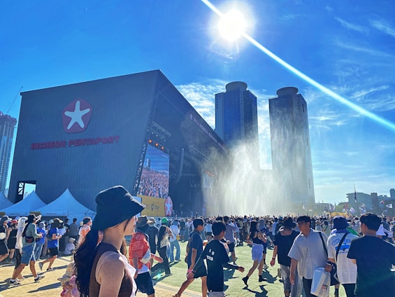 A large outdoor event with many people gathering in front of a stage. The stage has a big screen and is part of a structure labeled 'INCHEON PENTAPORT'. There are two prominent high-rise buildings in the background and the sun is shining brightly overhead with a clear blue sky. Some people are walking around wearing hats, and there are tents and flags visible in the surroundings. Water appears to be spraying in the air near the stage, possibly as a cooling mechanism for the crowd.