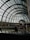 A grand indoor shopping environment with a large arched glass ceiling allowing natural light to fill the space. The architecture features intricate metal frameworks and symmetrical design. People are walking along a polished floor with various stores on either side. Prominent brand logos are visible, and there is decorative indoor greenery.