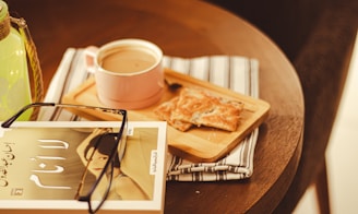 A wooden table with a striped cloth holds a cup of coffee in a pink mug on a wooden tray, accompanied by pastries. A book featuring an Arabic cover and a pair of glasses rests beside them. A green glass bottle is partly visible on the left side.