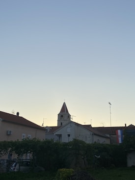 A charming landscape featuring a church with a tall, red-roofed bell tower surrounded by buildings with red-tiled roofs. The scene is set during dusk or dawn, as indicated by the gentle gradient of the sky, transitioning from a pale yellow near the horizon to a soft blue. Verdant greenery occupies the foreground, while a Croatian flag is visible on one of the buildings.