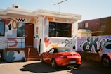 A small, single-story building with a colorful mural covering its walls. The artwork includes abstract shapes and playful figures in various colors. A red convertible car is parked in front, with a purple graffiti-covered fence and another vehicle visible to the right.