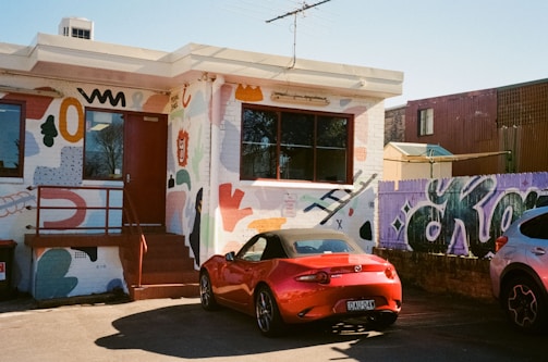 A small, single-story building with a colorful mural covering its walls. The artwork includes abstract shapes and playful figures in various colors. A red convertible car is parked in front, with a purple graffiti-covered fence and another vehicle visible to the right.