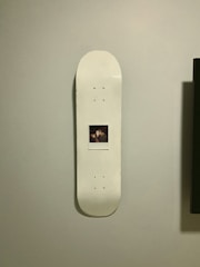 A skateboard deck with a white surface is mounted vertically on a wall. In the center of the skateboard is a Polaroid photograph featuring two people.