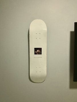 A skateboard deck with a white surface is mounted vertically on a wall. In the center of the skateboard is a Polaroid photograph featuring two people.
