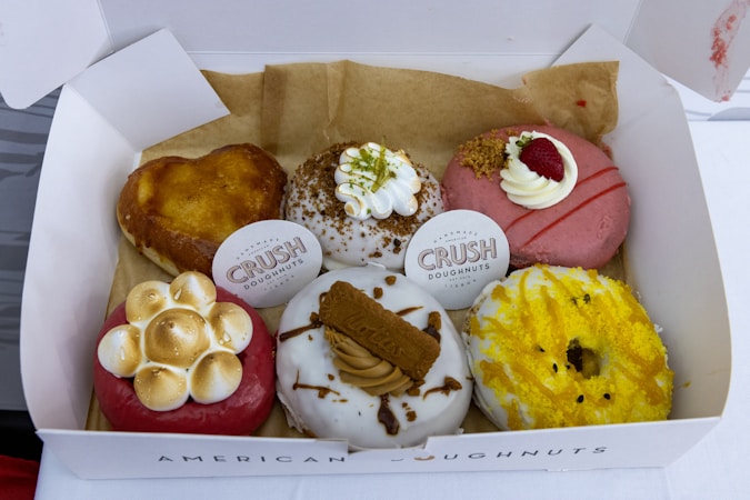 A box containing six assorted gourmet doughnuts, each with a unique topping and design. The doughnuts include a heart-shaped one, another covered with toasted marshmallow blobs, and a pink doughnut with cream and a raspberry on top. There are doughnuts garnished with yellow sprinkles, crushed nuts, and a cookie piece, each resting on a piece of brown parchment paper inside the box.