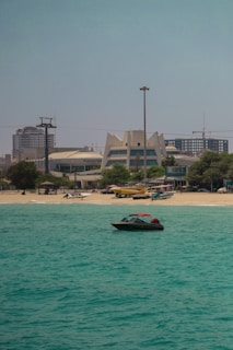 A small motorboat with a red roof is floating on turquoise water near a sandy beach. In the background, there are unique modern buildings with geometric architecture, surrounded by some trees. A few other small boats are parked on the shore, and a cityscape with high-rise buildings can be seen in the distance.