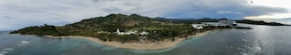 A panoramic view of a tropical coastline with a lush green landscape and rolling hills in the background. The scene includes a sandy beach, palm trees, and clear blue waters. A large cruise ship is visible docked near the coast, adding a touch of modernity to the natural setting.