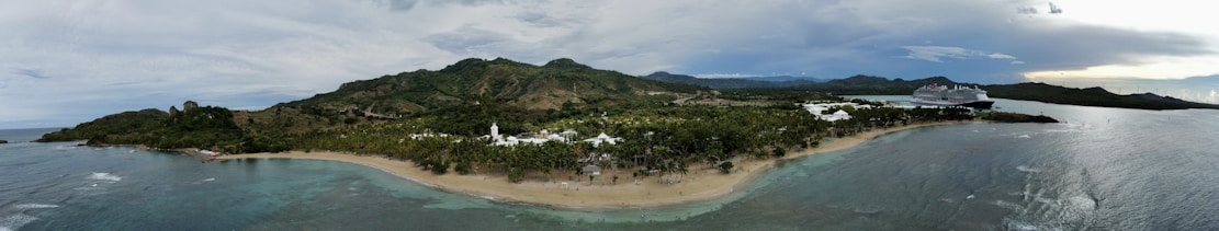 A panoramic view of a tropical coastline with a lush green landscape and rolling hills in the background. The scene includes a sandy beach, palm trees, and clear blue waters. A large cruise ship is visible docked near the coast, adding a touch of modernity to the natural setting.