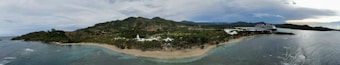 A panoramic view of a tropical coastline with a lush green landscape and rolling hills in the background. The scene includes a sandy beach, palm trees, and clear blue waters. A large cruise ship is visible docked near the coast, adding a touch of modernity to the natural setting.