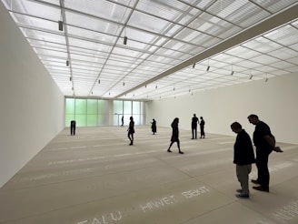 A spacious art gallery with a high, translucent ceiling allowing natural light to bathe the room. The floor features text inscriptions, and several people are scattered throughout the space, observing the environment. The walls are minimally decorated, enhancing the room's open and modern aesthetic.