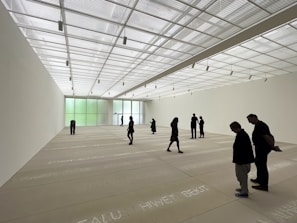 A spacious art gallery with a high, translucent ceiling allowing natural light to bathe the room. The floor features text inscriptions, and several people are scattered throughout the space, observing the environment. The walls are minimally decorated, enhancing the room's open and modern aesthetic.