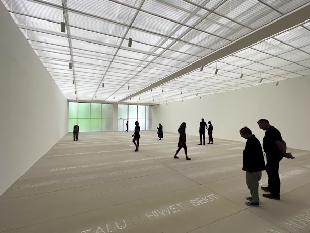 A spacious art gallery with a high, translucent ceiling allowing natural light to bathe the room. The floor features text inscriptions, and several people are scattered throughout the space, observing the environment. The walls are minimally decorated, enhancing the room's open and modern aesthetic.