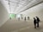 A spacious art gallery with a high, translucent ceiling allowing natural light to bathe the room. The floor features text inscriptions, and several people are scattered throughout the space, observing the environment. The walls are minimally decorated, enhancing the room's open and modern aesthetic.