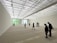 A spacious art gallery with a high, translucent ceiling allowing natural light to bathe the room. The floor features text inscriptions, and several people are scattered throughout the space, observing the environment. The walls are minimally decorated, enhancing the room's open and modern aesthetic.