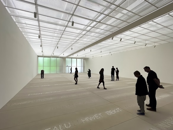 A spacious art gallery with a high, translucent ceiling allowing natural light to bathe the room. The floor features text inscriptions, and several people are scattered throughout the space, observing the environment. The walls are minimally decorated, enhancing the room's open and modern aesthetic.