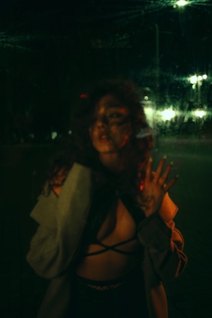 A mysterious figure stands in dim lighting, partially obscured by a reflective surface. The ambient glow from distant streetlights casts subtle highlights, and the person's hand is pressed against the glass. The attire includes a strappy top and a coat slipping from the shoulders, suggesting a casual yet dramatic pose.