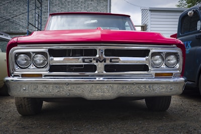 A vintage red GMC truck is parked on a gravel surface. The truck's front grille and headlights are prominently displayed, with shiny chrome detailing reflecting light. The background includes a brick building and parts of other vehicles.