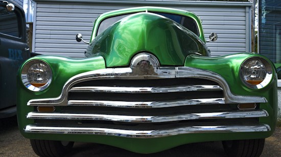 A vintage car with a shiny chrome grille and vibrant green paint. The car's rounded headlights and sleek design suggest a classic automobile aesthetic. It is parked in front of a corrugated metal garage with the shadow of another vehicle visible to the side.