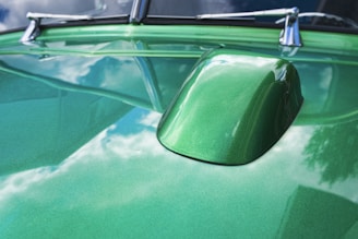 A shiny, green car hood with a raised section reflects a partly cloudy sky. The surface has a glossy finish, and there is a reflection of clouds and trees in the paint.