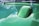 A shiny, green car hood with a raised section reflects a partly cloudy sky. The surface has a glossy finish, and there is a reflection of clouds and trees in the paint.