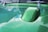 A shiny, green car hood with a raised section reflects a partly cloudy sky. The surface has a glossy finish, and there is a reflection of clouds and trees in the paint.
