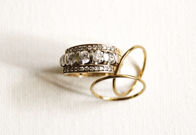 A pair of gold rings is lying on a white surface. One of the rings is adorned with several large, clear gemstones, possibly diamonds, set in a double row of smaller gemstones.