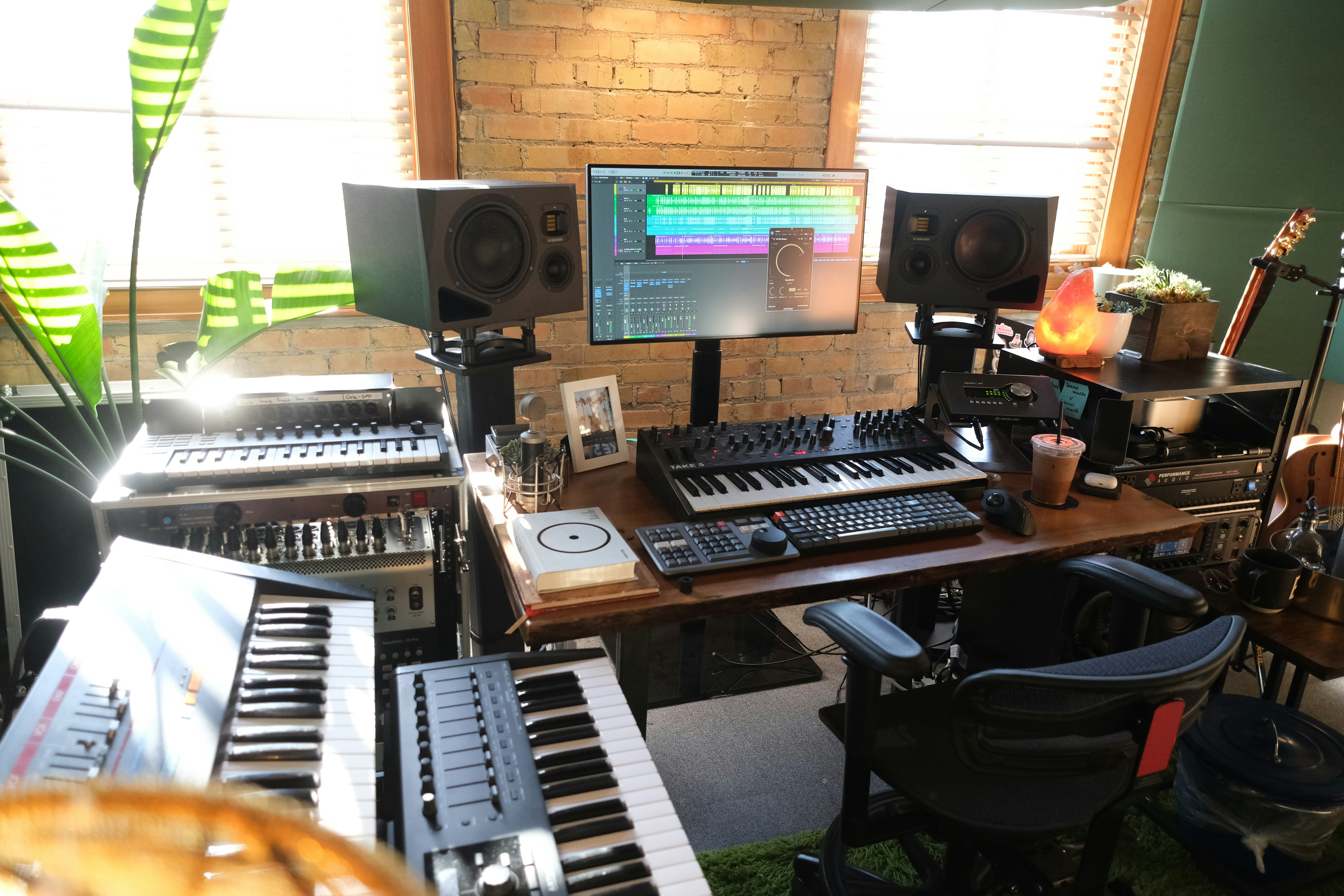 A home music studio featuring multiple electronic keyboards, a large monitor displaying audio editing software, and studio speakers. The room has a cozy vibe with a brick wall, wooden desk, potted plant, and a salt lamp. Various musical and audio equipment are neatly arranged, including synthesizers and mixers, alongside a comfortable office chair.