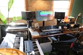 A home music studio featuring multiple electronic keyboards, a large monitor displaying audio editing software, and studio speakers. The room has a cozy vibe with a brick wall, wooden desk, potted plant, and a salt lamp. Various musical and audio equipment are neatly arranged, including synthesizers and mixers, alongside a comfortable office chair.