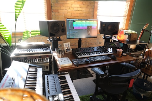 A home music studio featuring multiple electronic keyboards, a large monitor displaying audio editing software, and studio speakers. The room has a cozy vibe with a brick wall, wooden desk, potted plant, and a salt lamp. Various musical and audio equipment are neatly arranged, including synthesizers and mixers, alongside a comfortable office chair.
