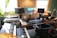 A home music studio featuring multiple electronic keyboards, a large monitor displaying audio editing software, and studio speakers. The room has a cozy vibe with a brick wall, wooden desk, potted plant, and a salt lamp. Various musical and audio equipment are neatly arranged, including synthesizers and mixers, alongside a comfortable office chair.