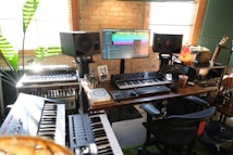 A home music studio featuring multiple electronic keyboards, a large monitor displaying audio editing software, and studio speakers. The room has a cozy vibe with a brick wall, wooden desk, potted plant, and a salt lamp. Various musical and audio equipment are neatly arranged, including synthesizers and mixers, alongside a comfortable office chair.